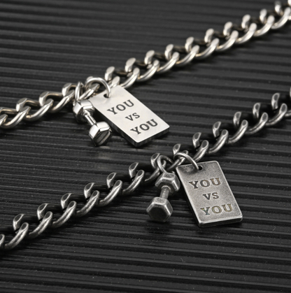 Silver chain necklace with 'YOU VS YOU' tag charms on a black textured background