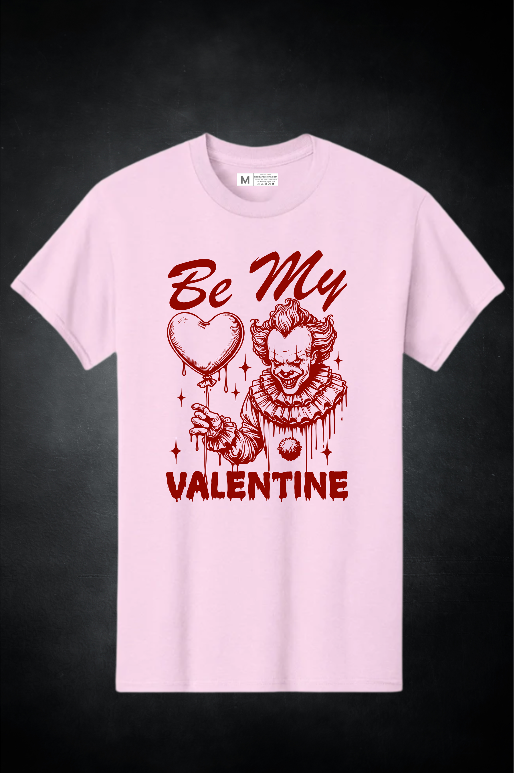 Pink t-shirt with a graphic design of a clown holding a heart and text 'Be My Valentine' on a dark background.