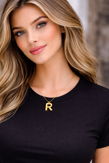 Woman wearing a black top and gold necklace with a 'R' pendant against a blurred background