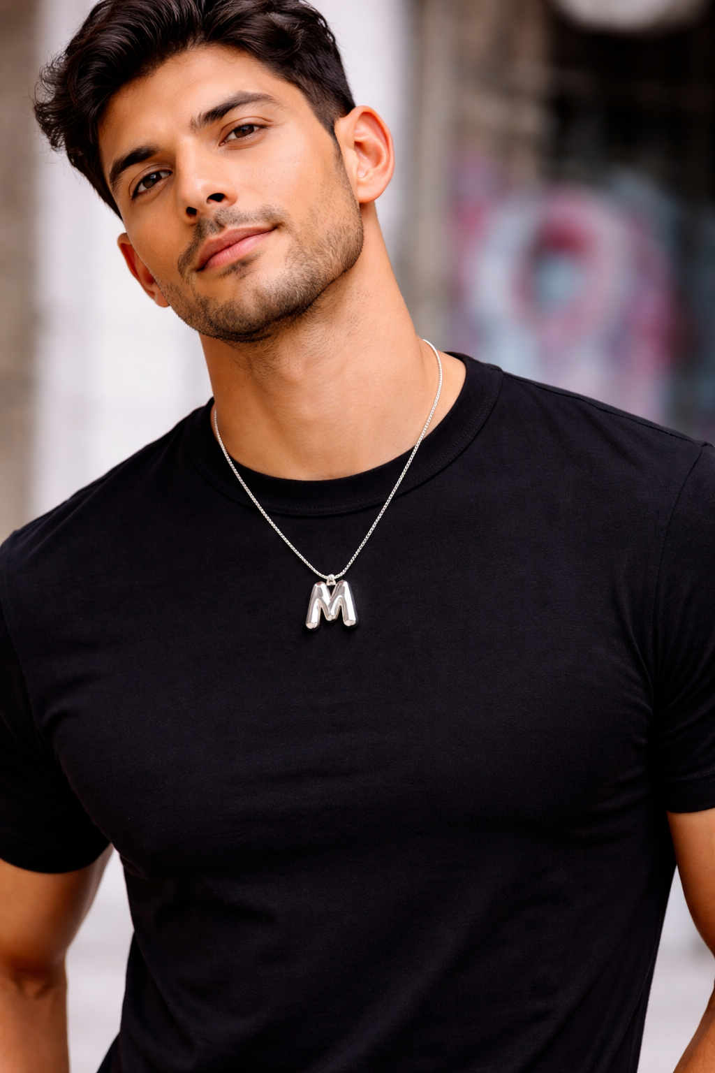 Man wearing a black t-shirt with a necklace featuring a 'M' pendant, standing against an urban background.
