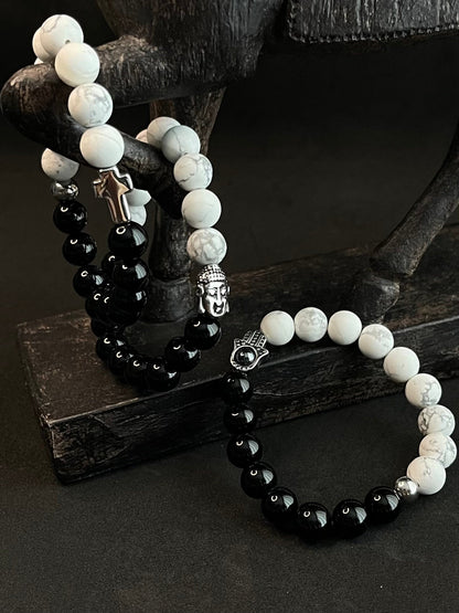 Black and white beaded bracelet set featuring glossy black onyx beads, white howlite stones, silver Buddha charm, cross pendant, and Hamsa hand charm displayed on a dark wooden stand.