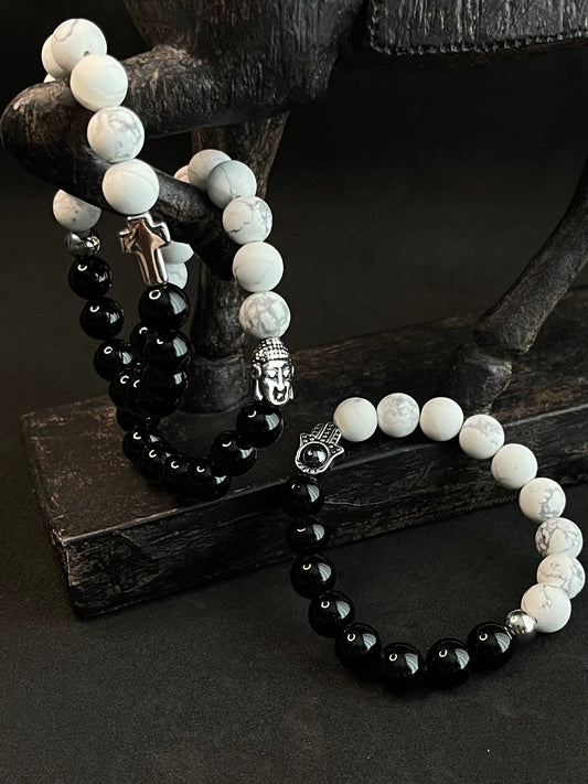 Black and white beaded bracelet set featuring glossy black onyx beads, white howlite stones, silver Buddha charm, cross pendant, and Hamsa hand charm displayed on a dark wooden stand.