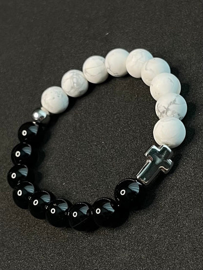 Men’s black obsidian and white howlite bead bracelet stack with silver Buddha charm, cross pendant, and Hamsa symbol on a rustic wooden display.