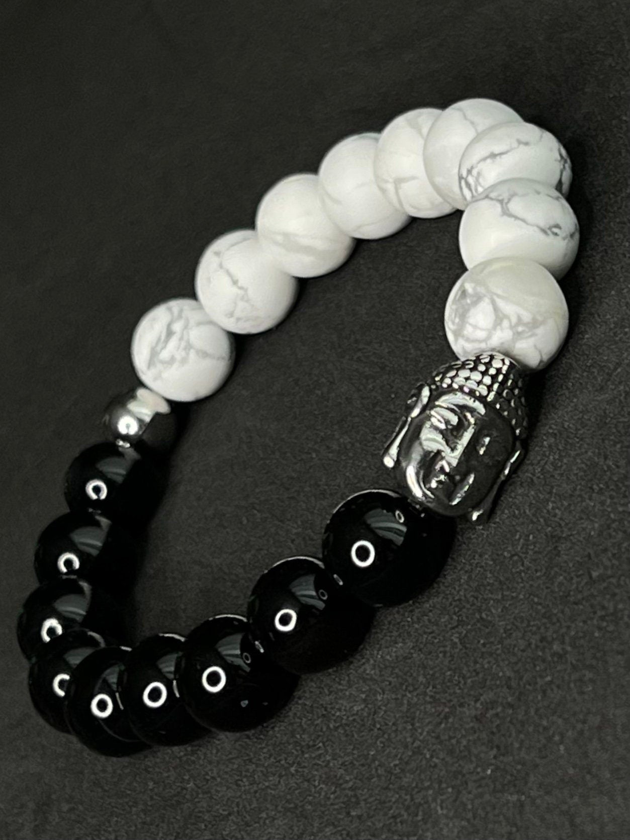 Spiritual black and white gemstone bracelet with polished black beads, white marbled howlite stones, silver Buddha head charm and symbolic cross and Hamsa accents.